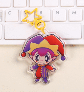 An Acrylic keychain with a yellow star clasp, placed on top of a white keyboard. The keychain features a small little clown with an alternating red/purple design. The clown is holding his arms out to the sides, looking very cute.