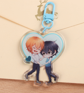 An Acrylic Keychain with a blue clasp sitting on a yellow envelope, featuring two characters holding hands. They're surrounded by a blue heart. The character on the left has orange hair and a big smile, the character on the right is shrunk down nervously and has black hair.